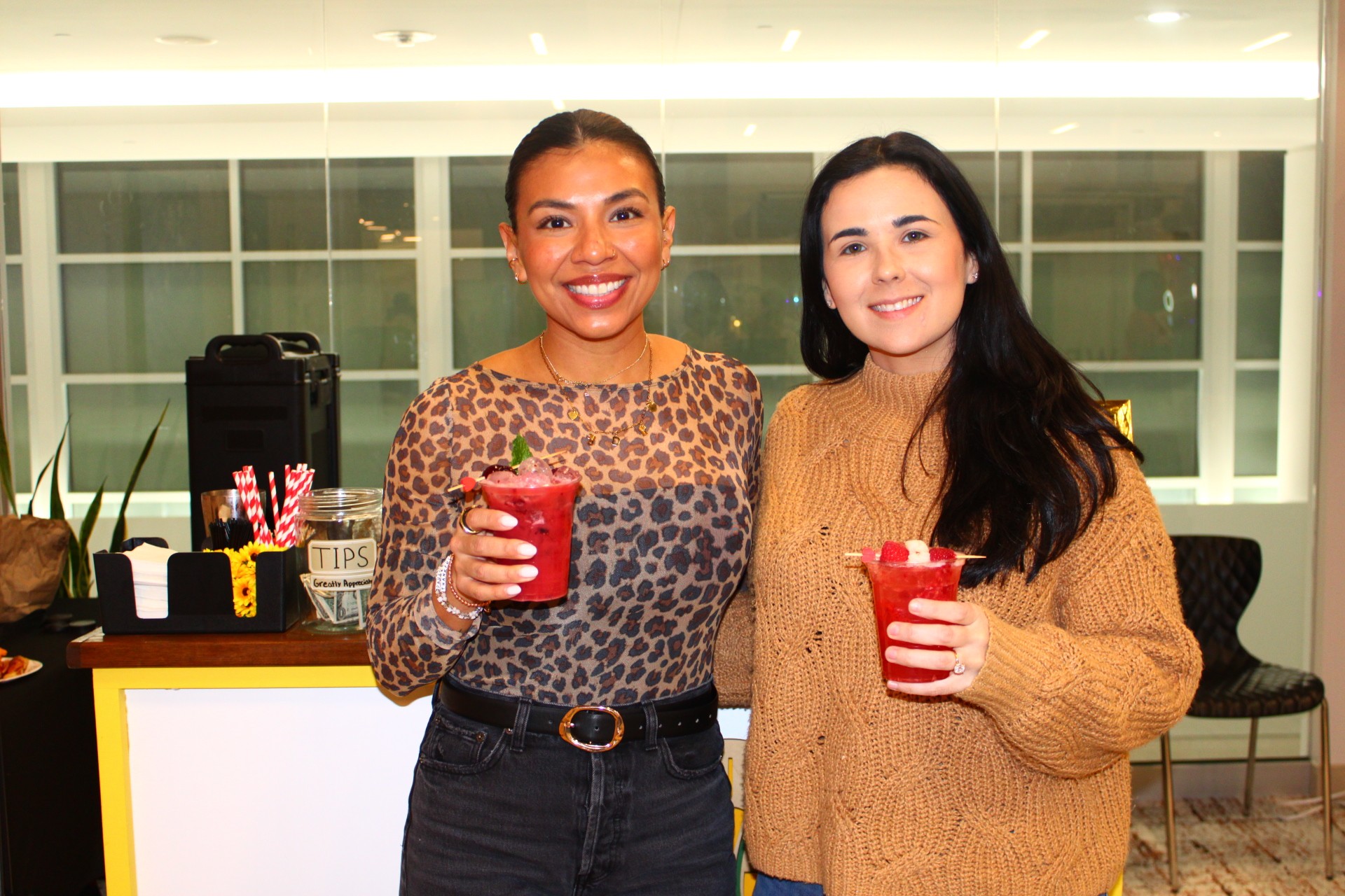 Two women smiling and holding red drinks indoors, with a window and chairs in the background.