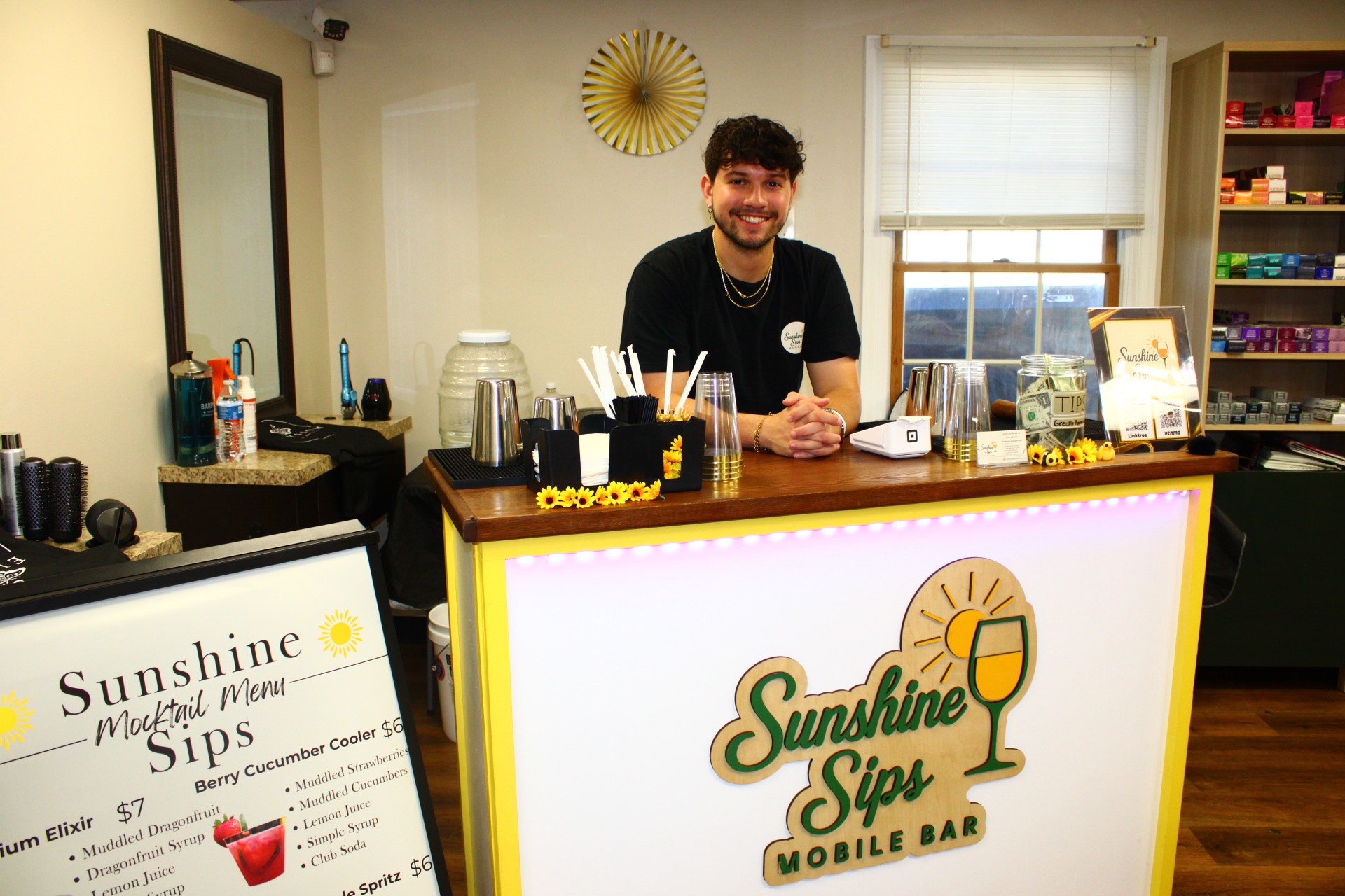 A smiling man stands behind a bar counter with a sign reading 'Sunshine Sip' and various drink supplies, in a cozy room.