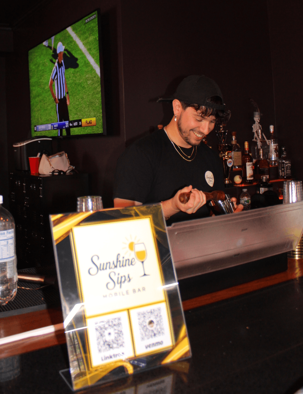 Person behind bar preparing a drink, bar setup with bottles, a TV screen, and a sign with QR codes.