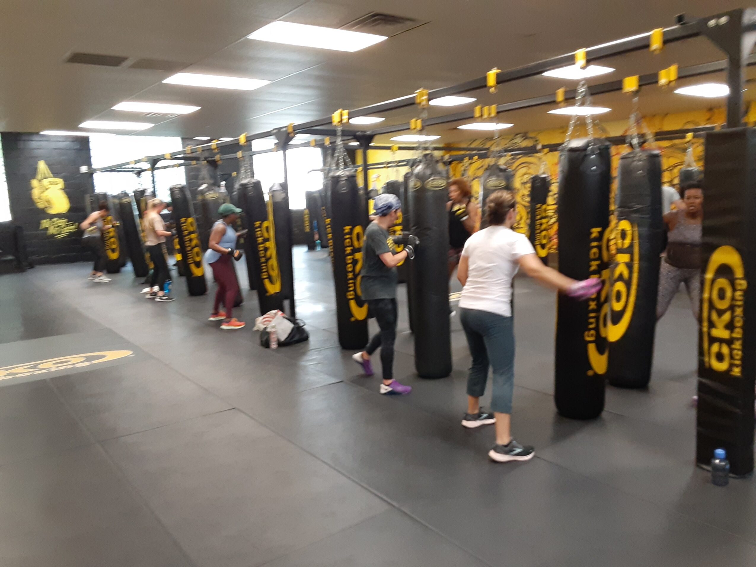 People practicing boxing with punching bags in a gym, some wearing gloves and athletic clothing, in a well-lit space.