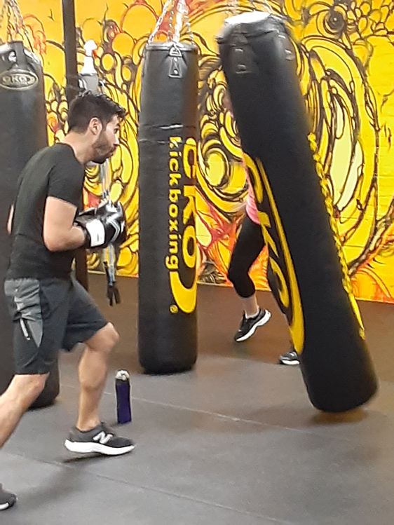 Two people practicing boxing with punching bags in a gym, one person in black shorts and gloves, the other behind a bag.