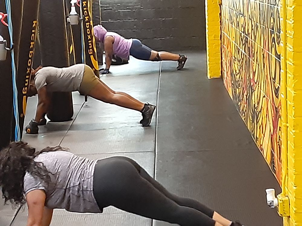 Three people stretching on the floor in a gym, with a colorful wall on the right side.