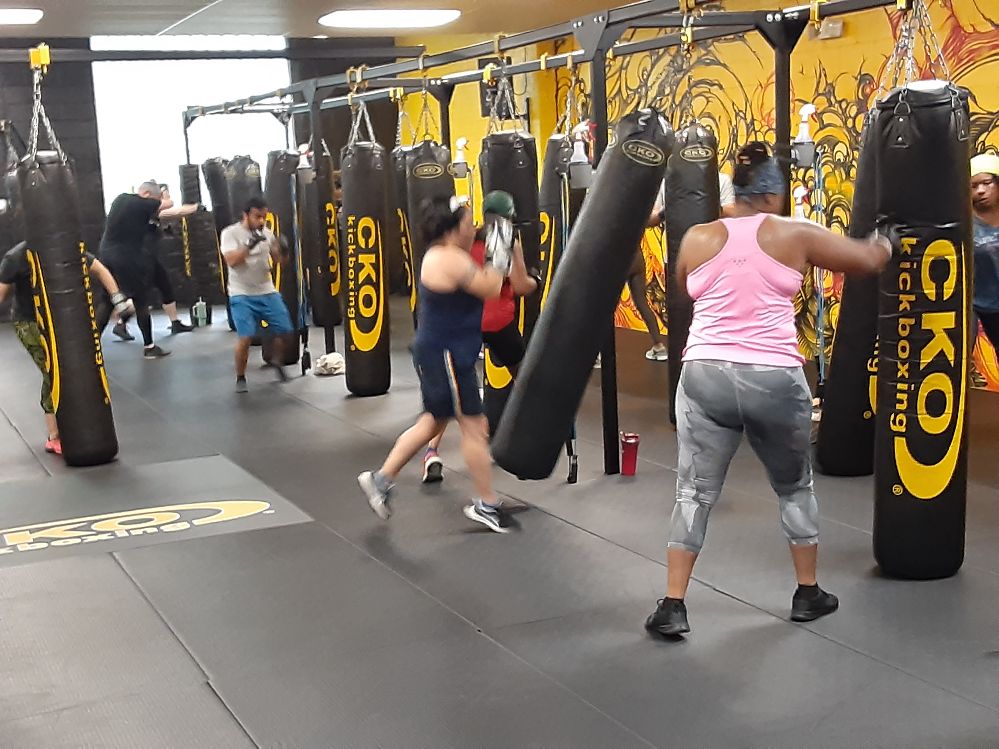 People practicing boxing with punching bags in a gym, some hitting bags, others preparing or observing, yellow and black decor.