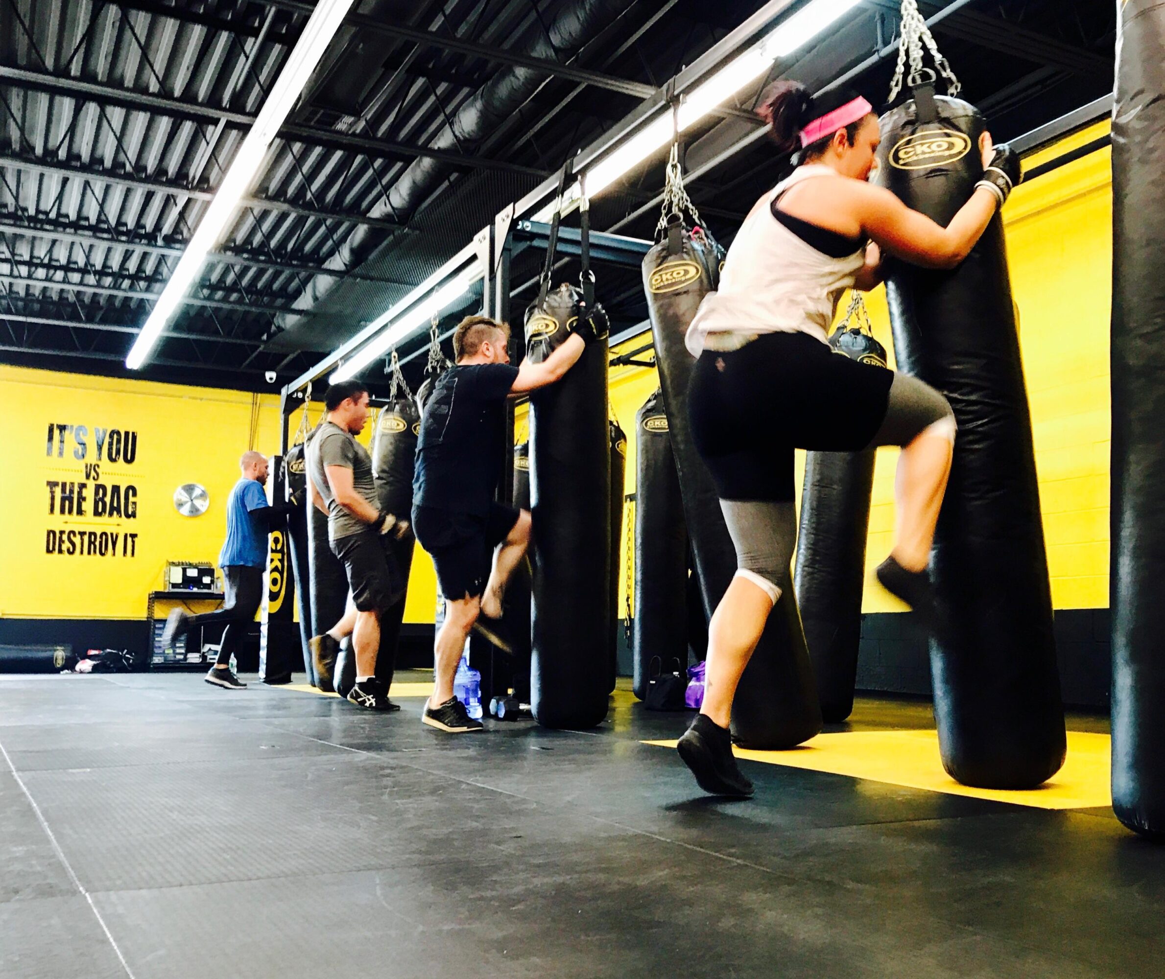 People exercising with punching bags in a gym, yellow and black walls, ceiling lights, and motivational text on the wall.