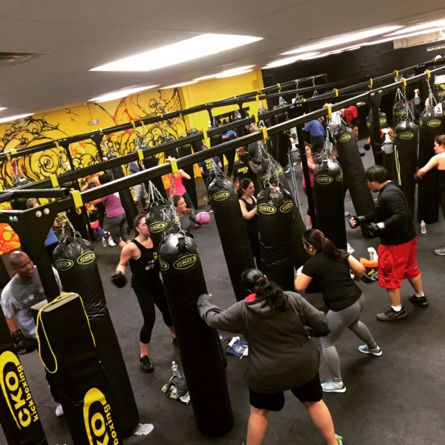 People practicing boxing in a gym with punching bags and a yellow wall mural, some wearing gloves and athletic clothing.