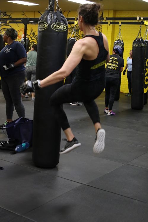 Woman in black workout clothes kicks a punching bag in a gym with other people training.