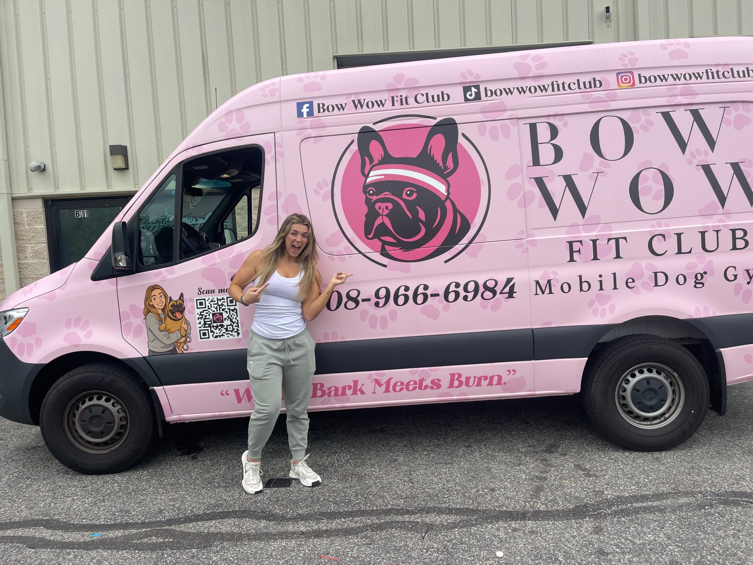 Woman standing next to a pink mobile dog grooming van with a French bulldog logo and text, outdoors.