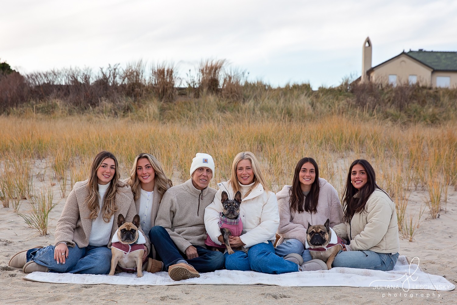 Group of six women sitting on a blanket outdoors with three French bulldogs, grassy field and lighthouse in background.