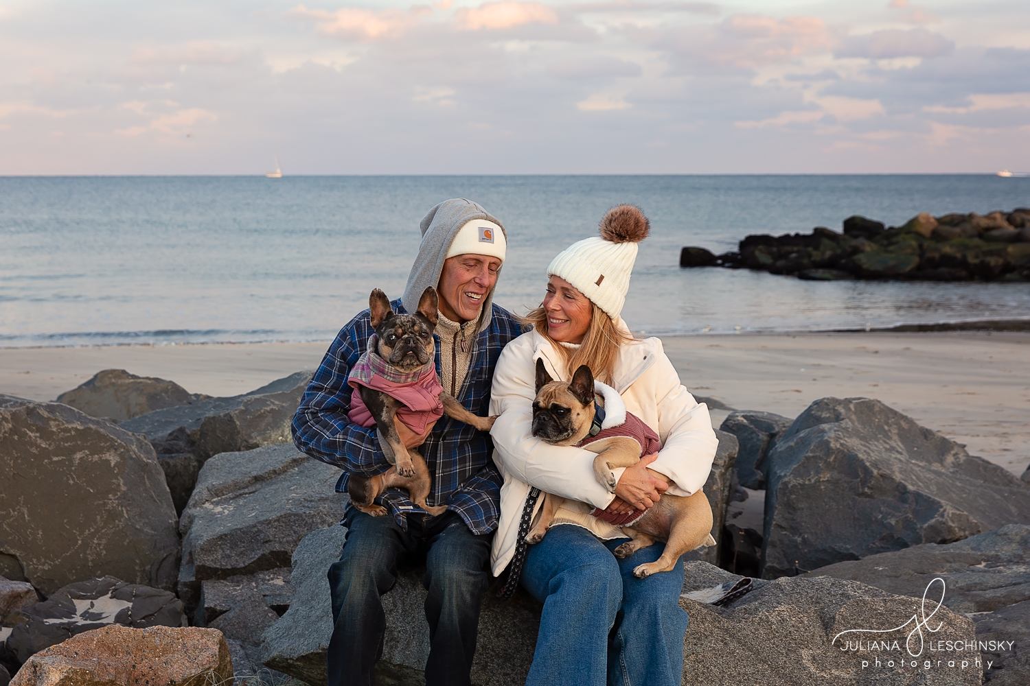A smiling couple with two dogs sitting on rocks by the ocean at sunset, wearing warm clothing and hats.