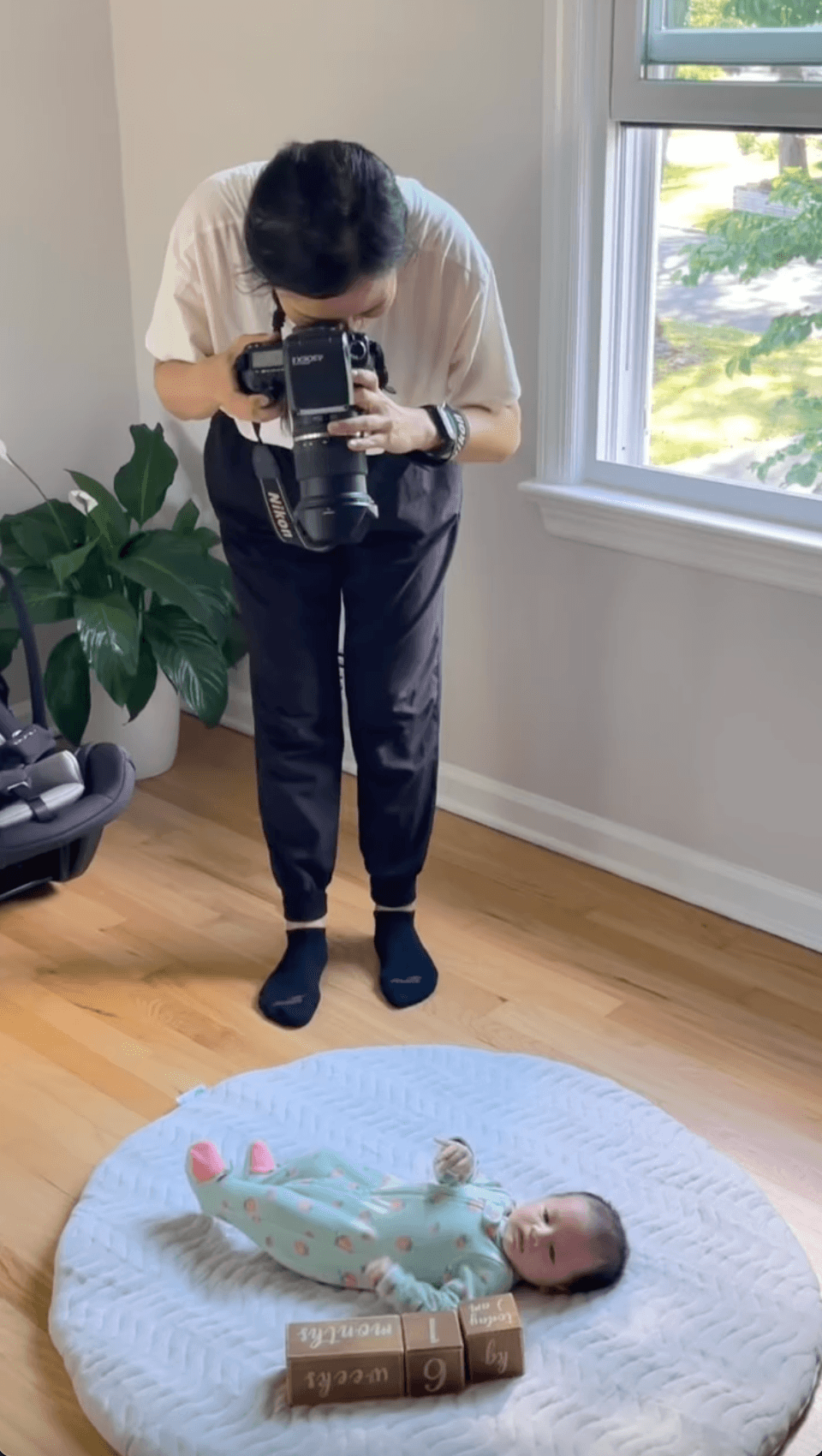 Person holding camera, looking down at a baby lying on a round rug near a window, with a plant nearby.