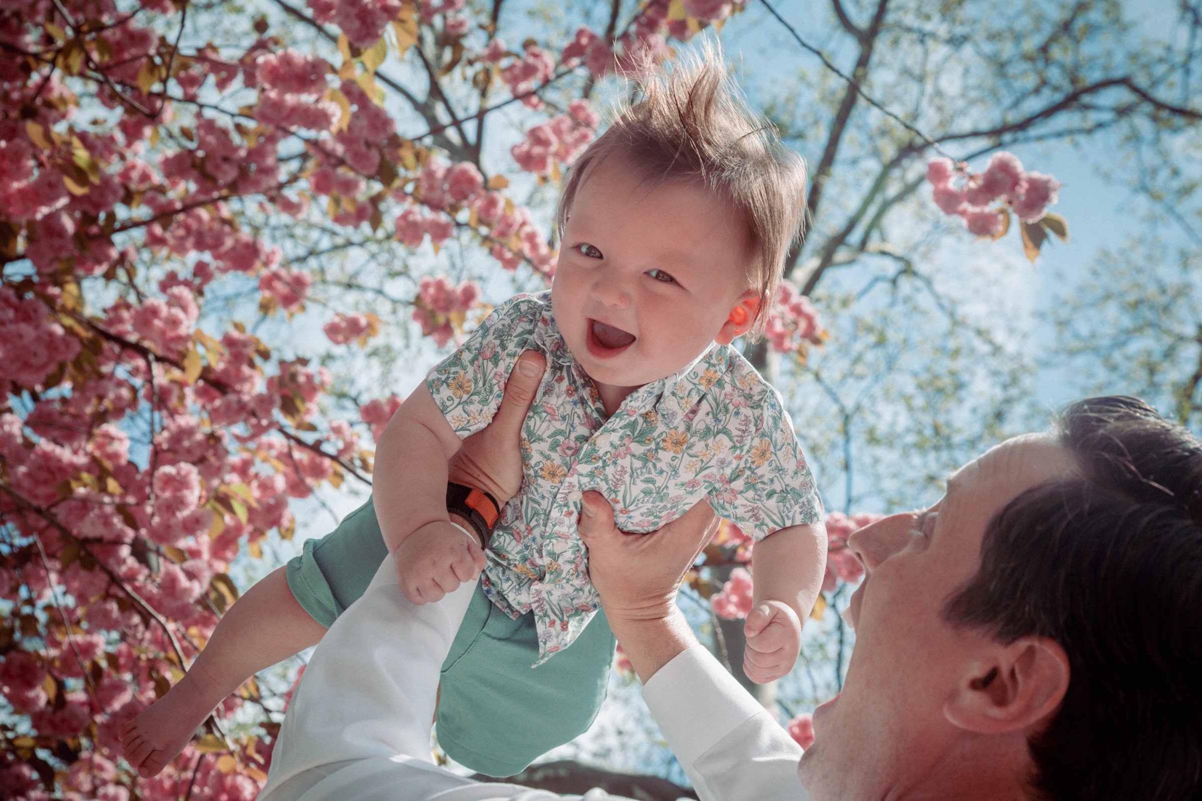 Child with spiky hair smiling and being lifted outdoors near pink blossoms and blue sky