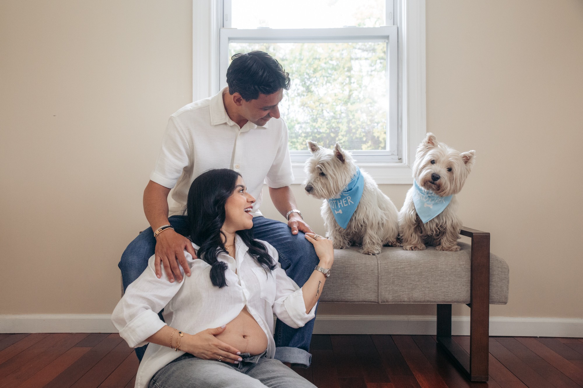 A family of three with two dogs sitting on a bench by a window in a room with beige walls and wooden floor.