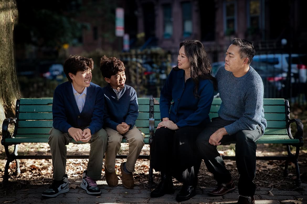 Four people sitting on a park bench, engaged in conversation, with trees and buildings in the background.