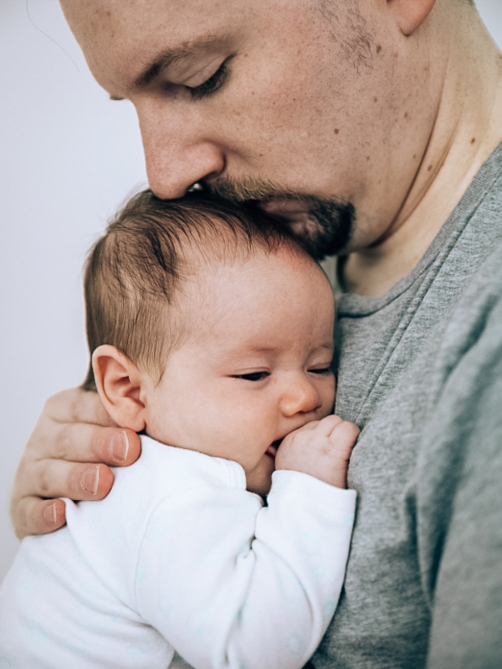 Adult holding a baby close, adult's face near baby's head, both with neutral expressions, plain background.
