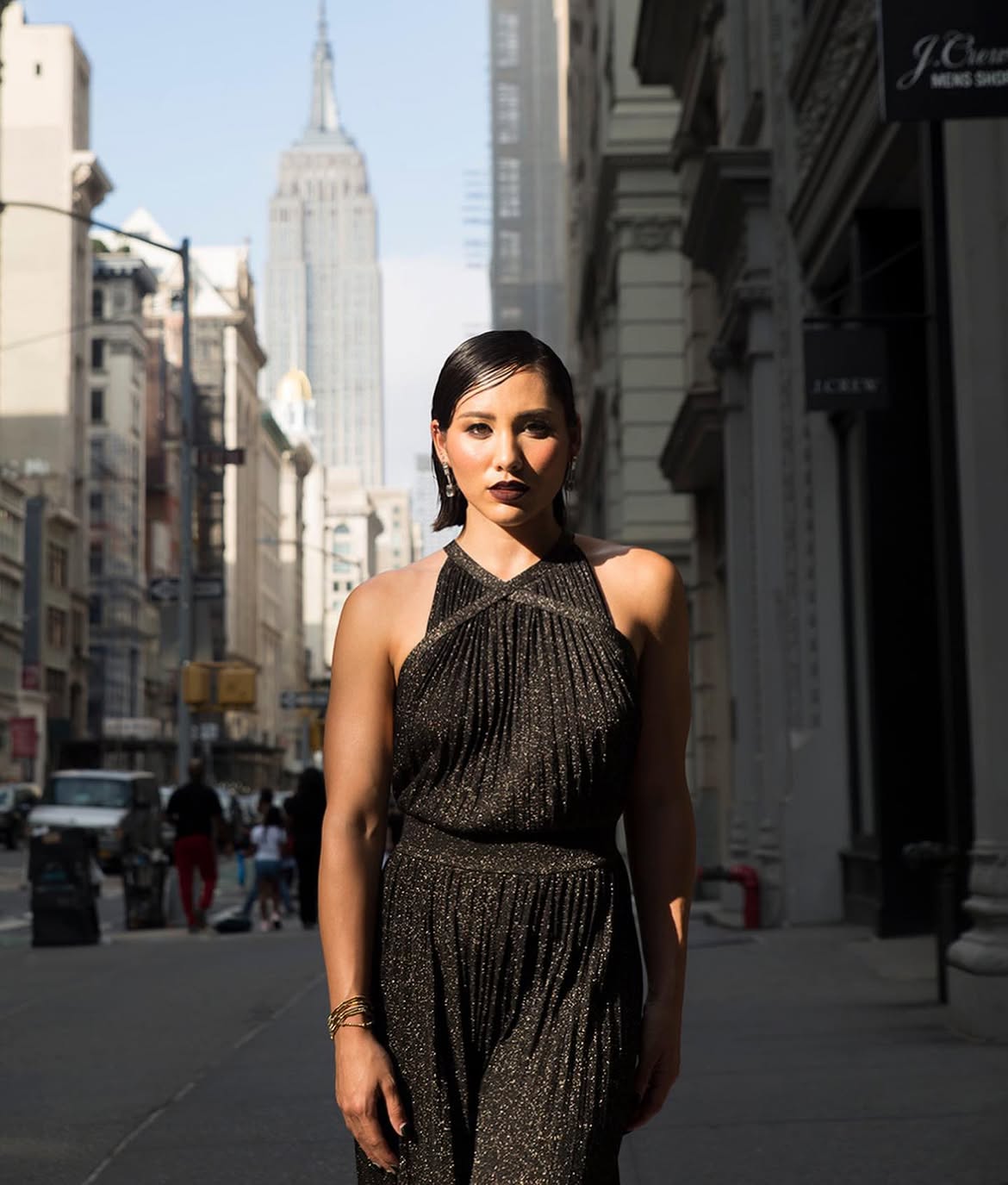 Woman in black dress standing on city street with tall buildings and Empire State Building in background.