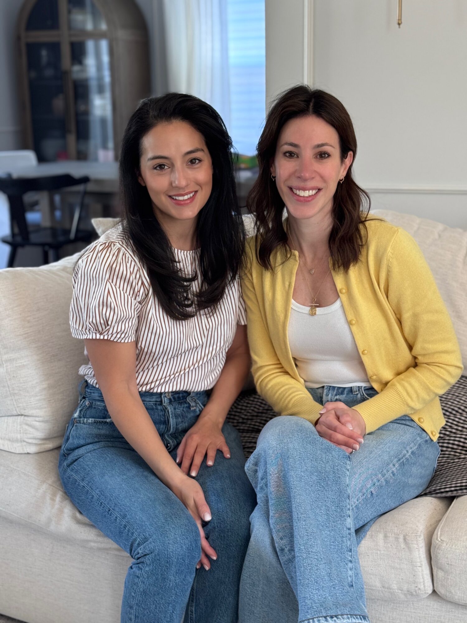 Two women sitting on a sofa, smiling, in a cozy room with a window and curtains behind them.