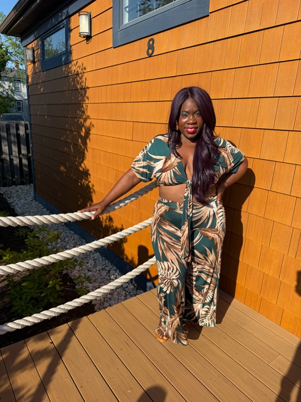 Woman with long dark hair in patterned outfit standing outside near a wooden wall with ropes, sunlight casting shadows.