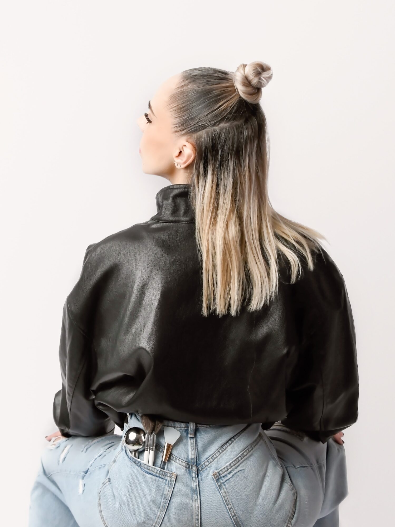 Woman with long hair in a bun, wearing a black satin blouse and light jeans, facing sideways against a white background.
