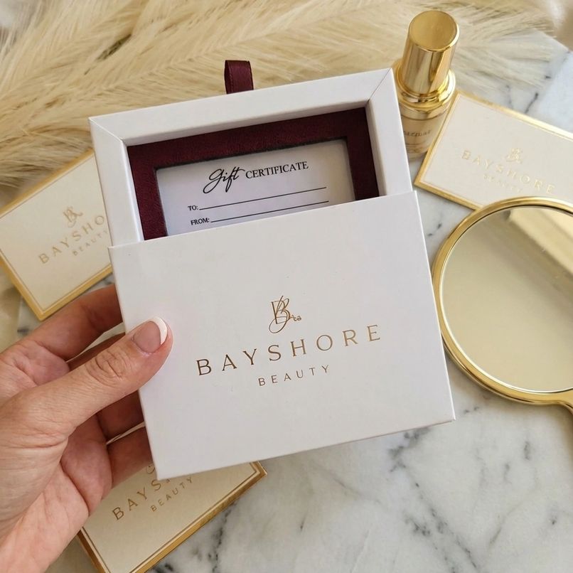 Hand holding a Bayshore Beauty gift certificate in a white box with gold lettering, on a marble surface with beauty products and a mirror.