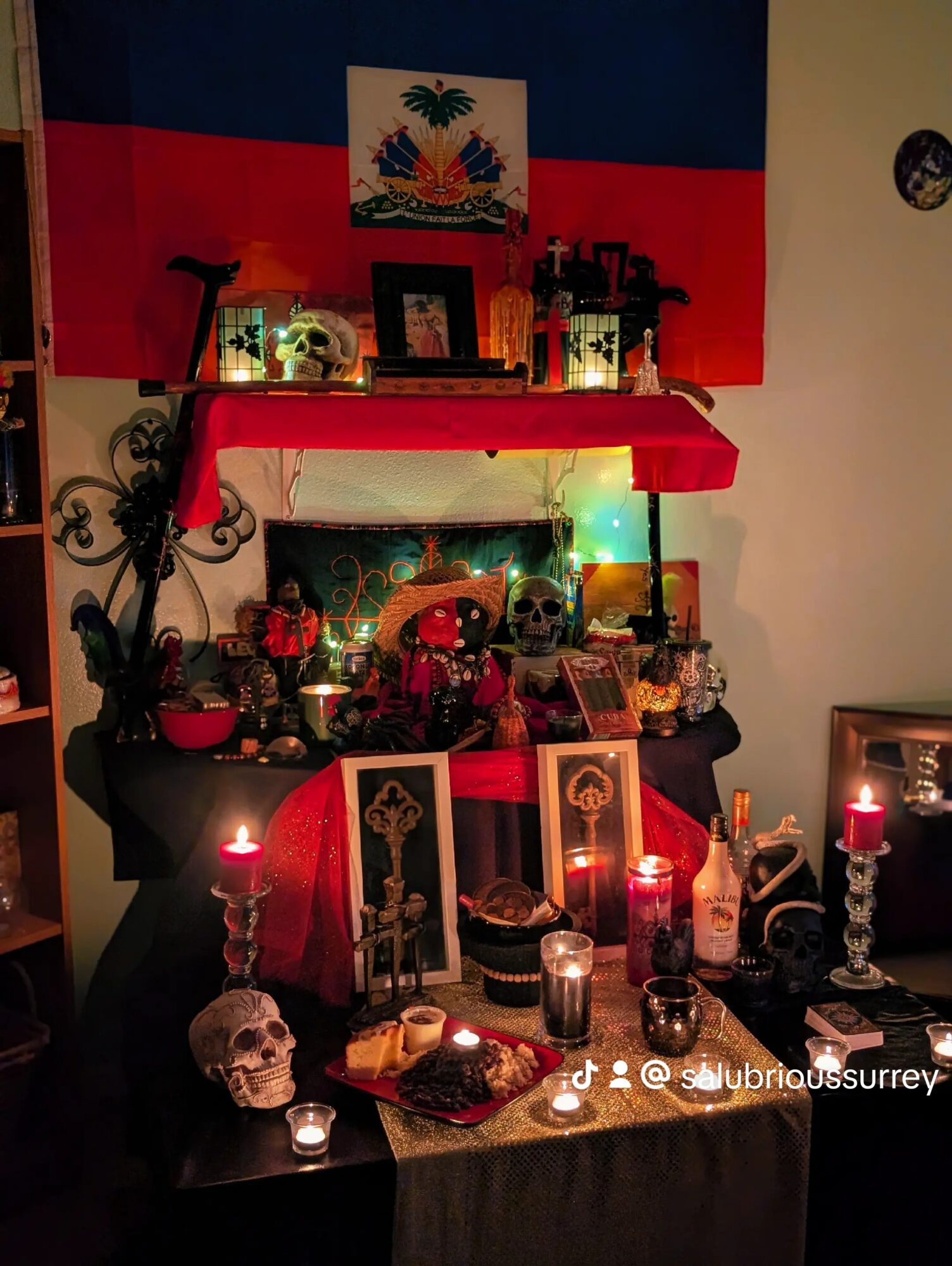 Altar with candles, skulls, and decorations, with a flag and picture above, in a dimly lit room.
