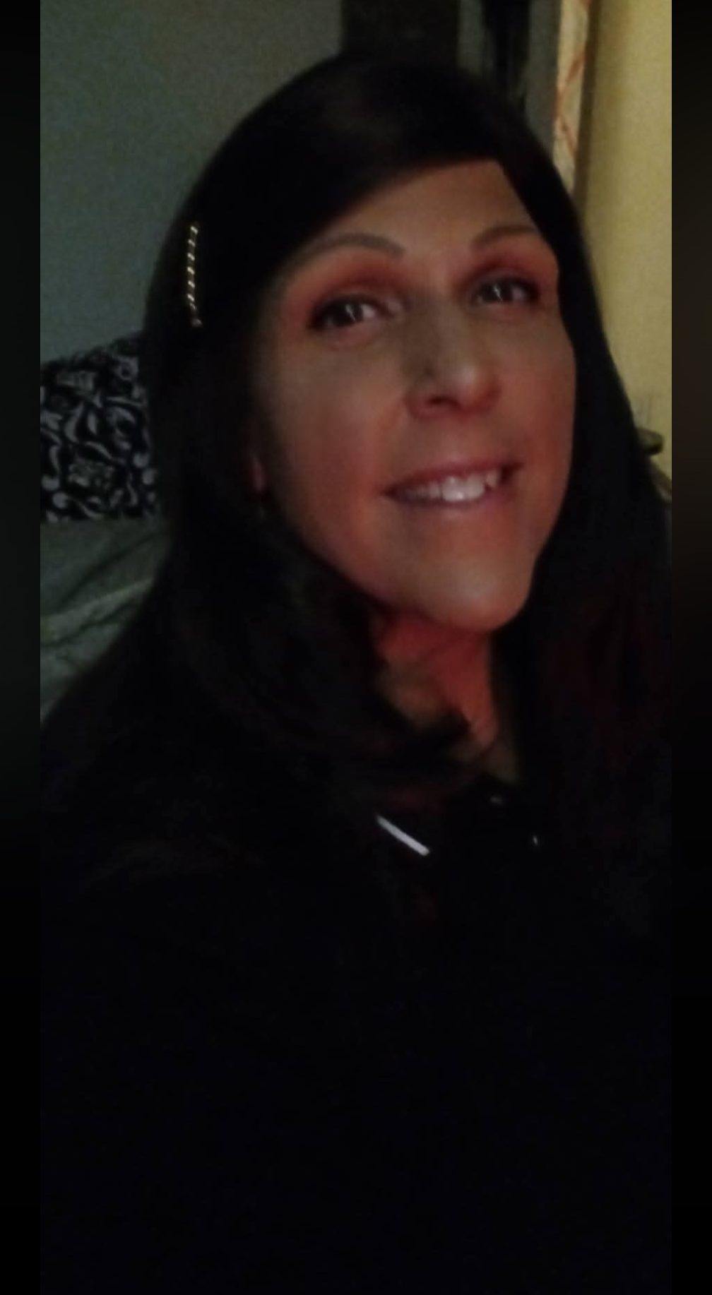 Woman with dark hair smiling, wearing a hair accessory, in a dimly lit indoor setting.