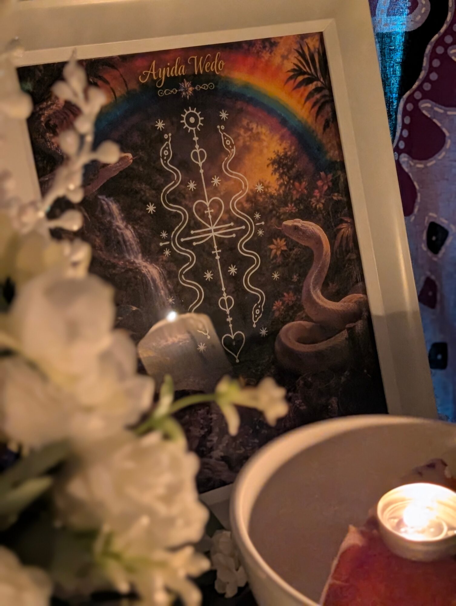 Framed colorful artwork with rainbow, snake, and decorative elements, partially obscured by white flowers and a lit candle.