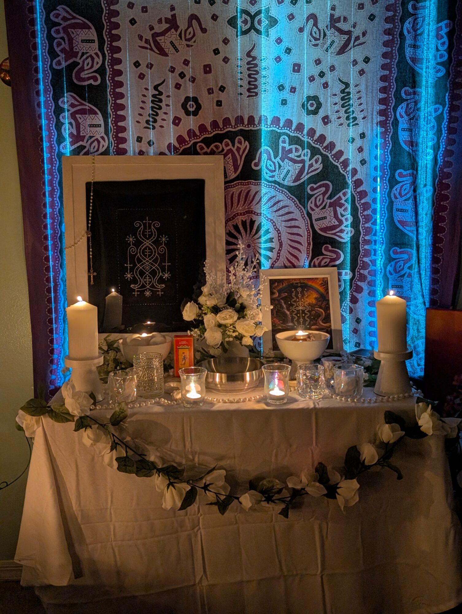 Table with candles, flowers, framed pictures, and a bowl, set against a colorful, patterned curtain backdrop.