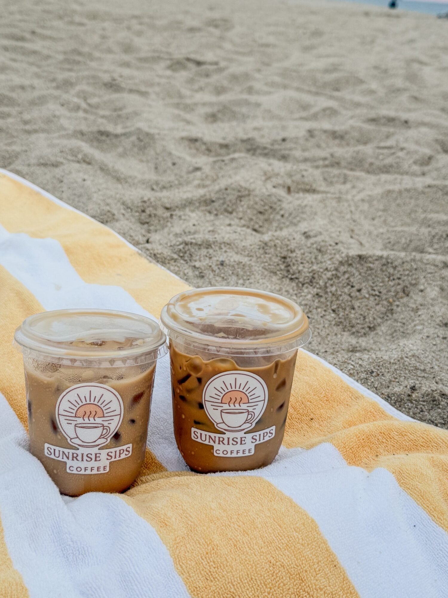 Two iced coffee drinks on a yellow and white striped towel at the beach, sand in the background.