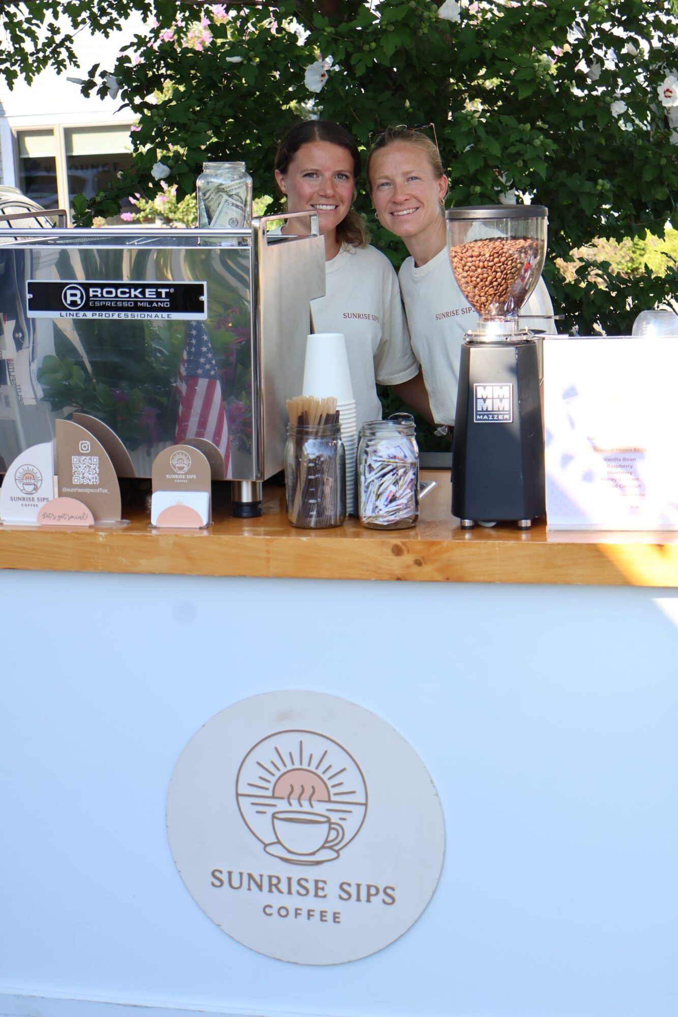 Coffee shop counter with cups, a teapot, and a woman smiling behind it, outdoors with trees and buildings in background.