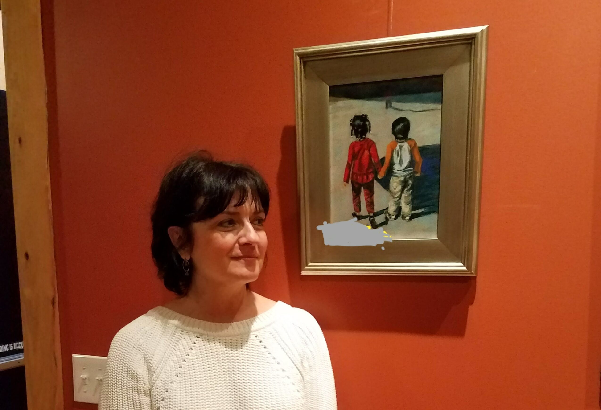 Woman with short dark hair wearing a white sweater standing in front of a red wall with framed painting of two children walking on a beach.