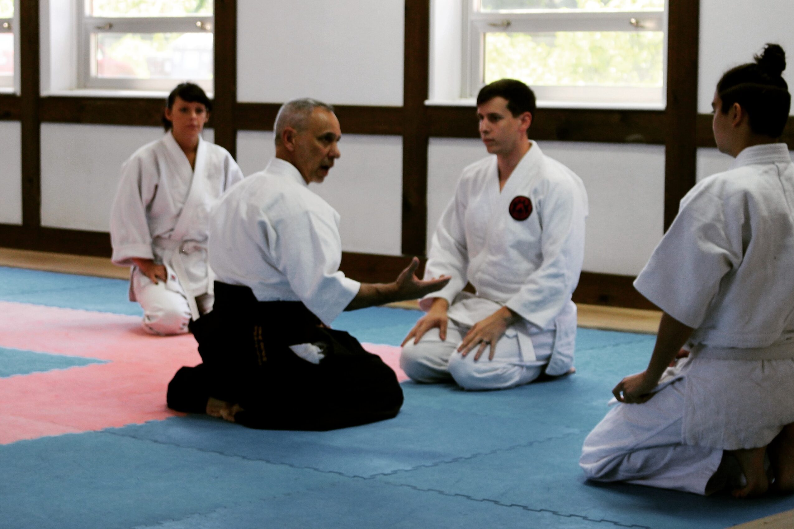 Four people in martial arts uniforms sit on mats in a dojo, with one person speaking to three others. Large windows in background.