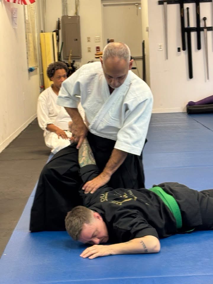 Instructor demonstrates a martial arts technique on a person lying on the floor, with another person observing in the background.