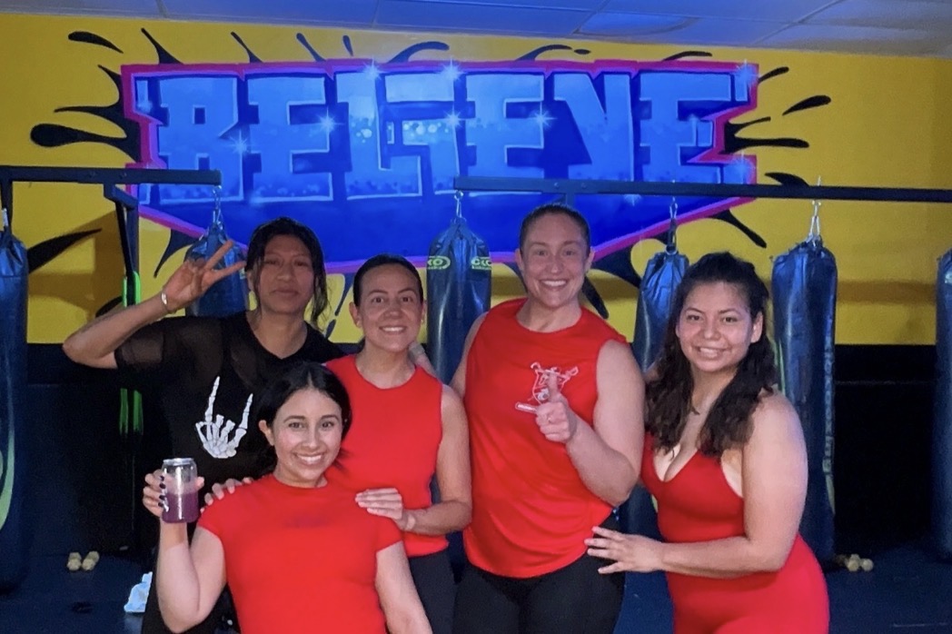 Five women in red and black workout clothes pose in a gym with punching bags and a colorful graffiti-style sign that says 'BELIEVE'.