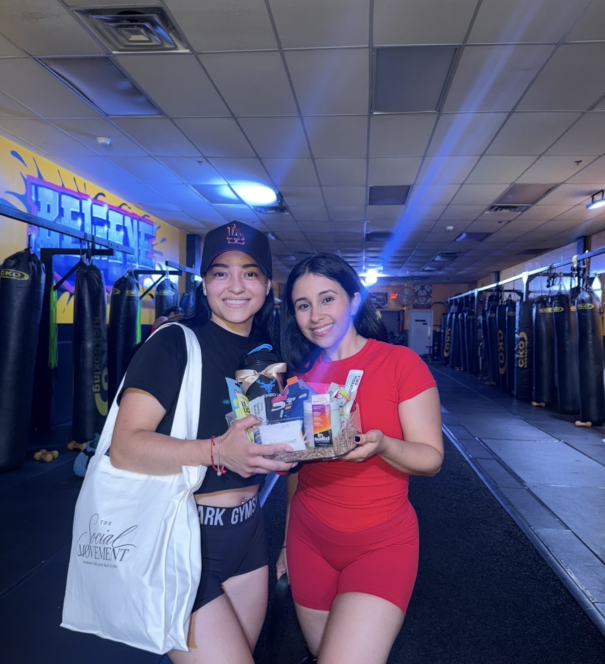 Two women smiling and holding a gift basket in a store with clothing racks, one in black and the other in red, under fluorescent lights.