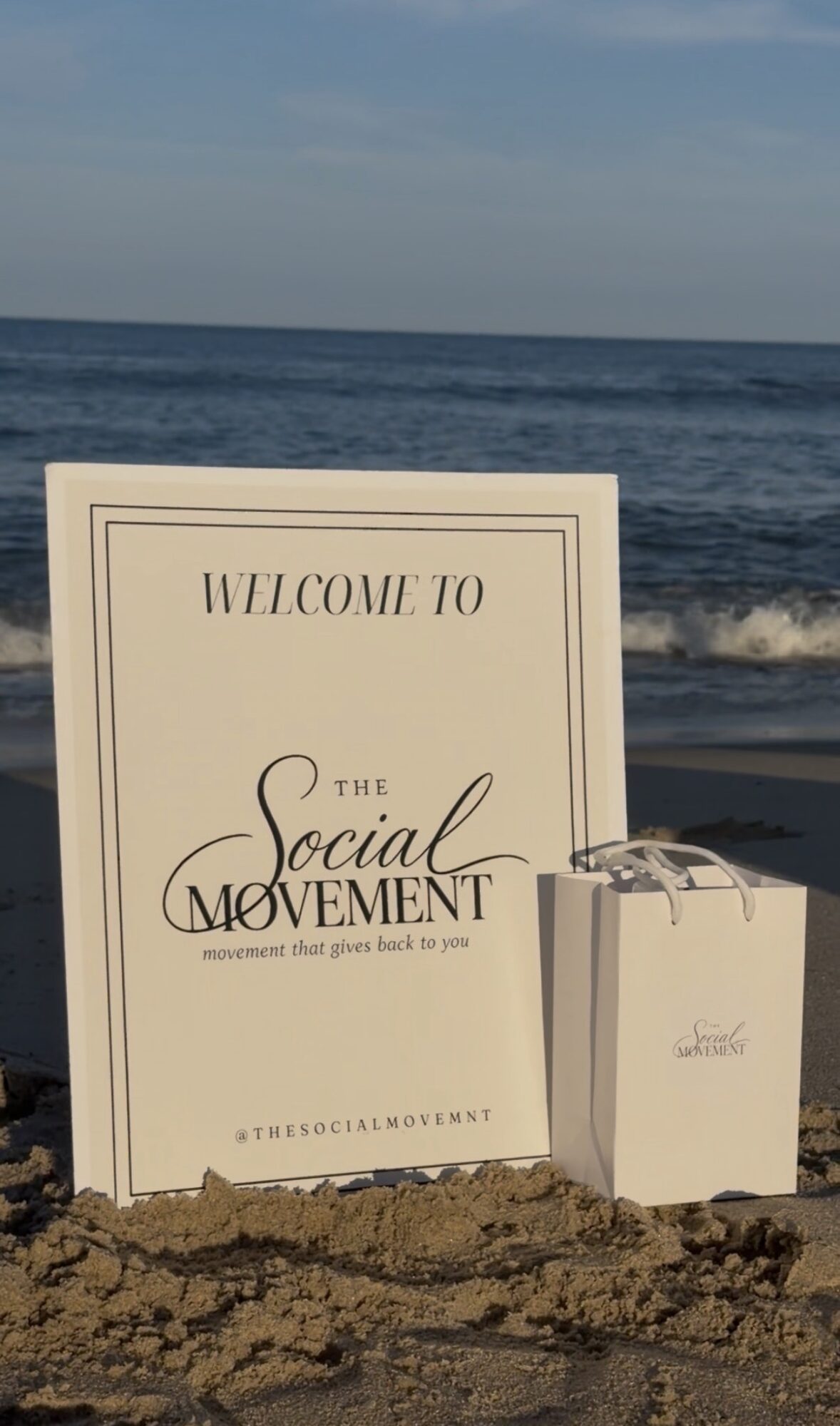 Sign on beach reads 'Welcome to the Social Movement' with a small white box beside it, ocean and sky in background.
