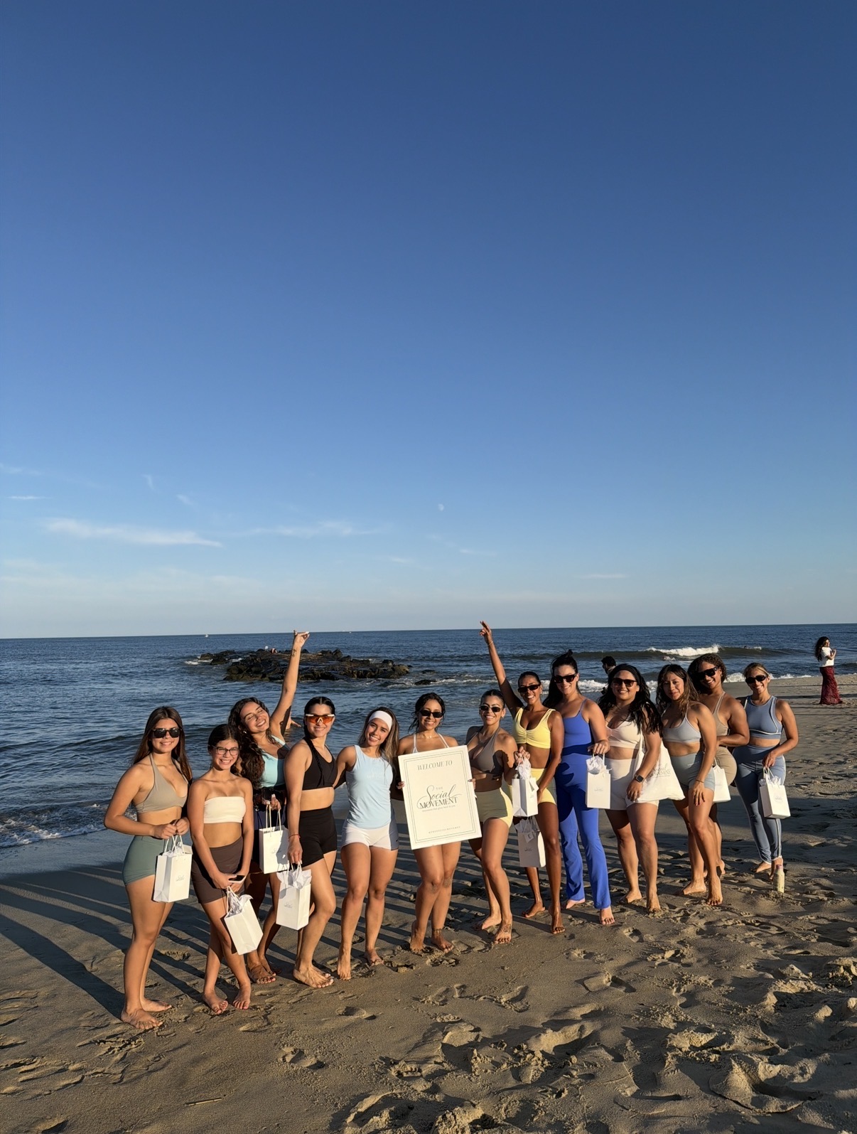 Group of people standing on a sandy beach near the ocean, holding signs, with a clear blue sky overhead.