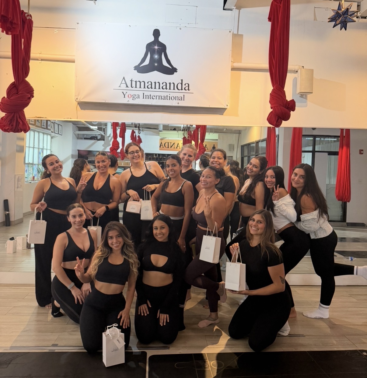 Group of women in yoga attire posing in a yoga studio with red curtains and a sign reading 'Atmandanda Yoga International'.