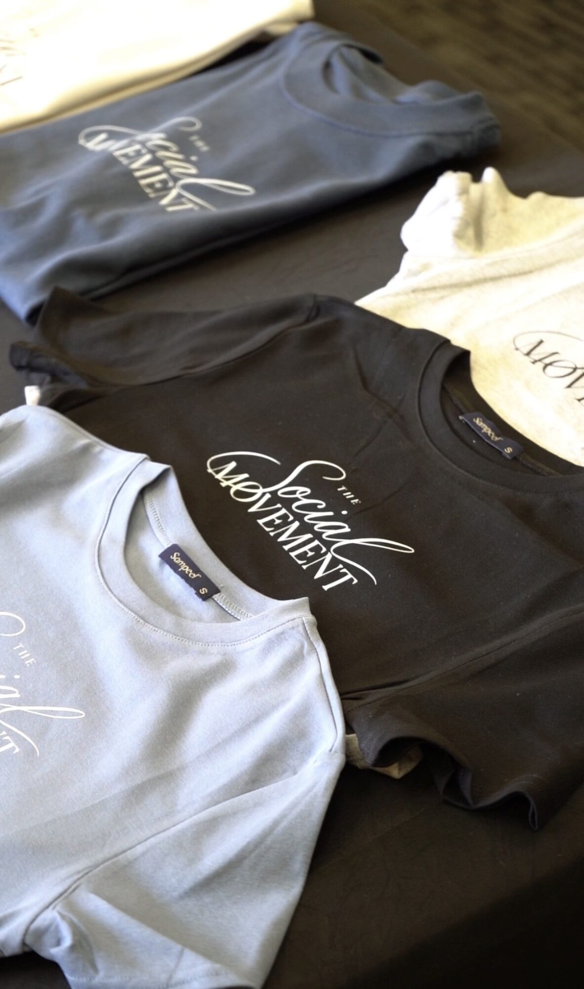 Four folded t-shirts in blue, black, white, and beige with printed text, laid on a dark surface.