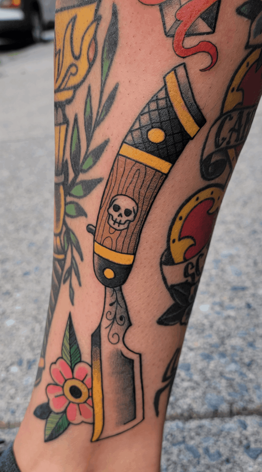 Tattoo of a wooden pistol with a skull, surrounded by colorful flowers and other tattoos on arm.