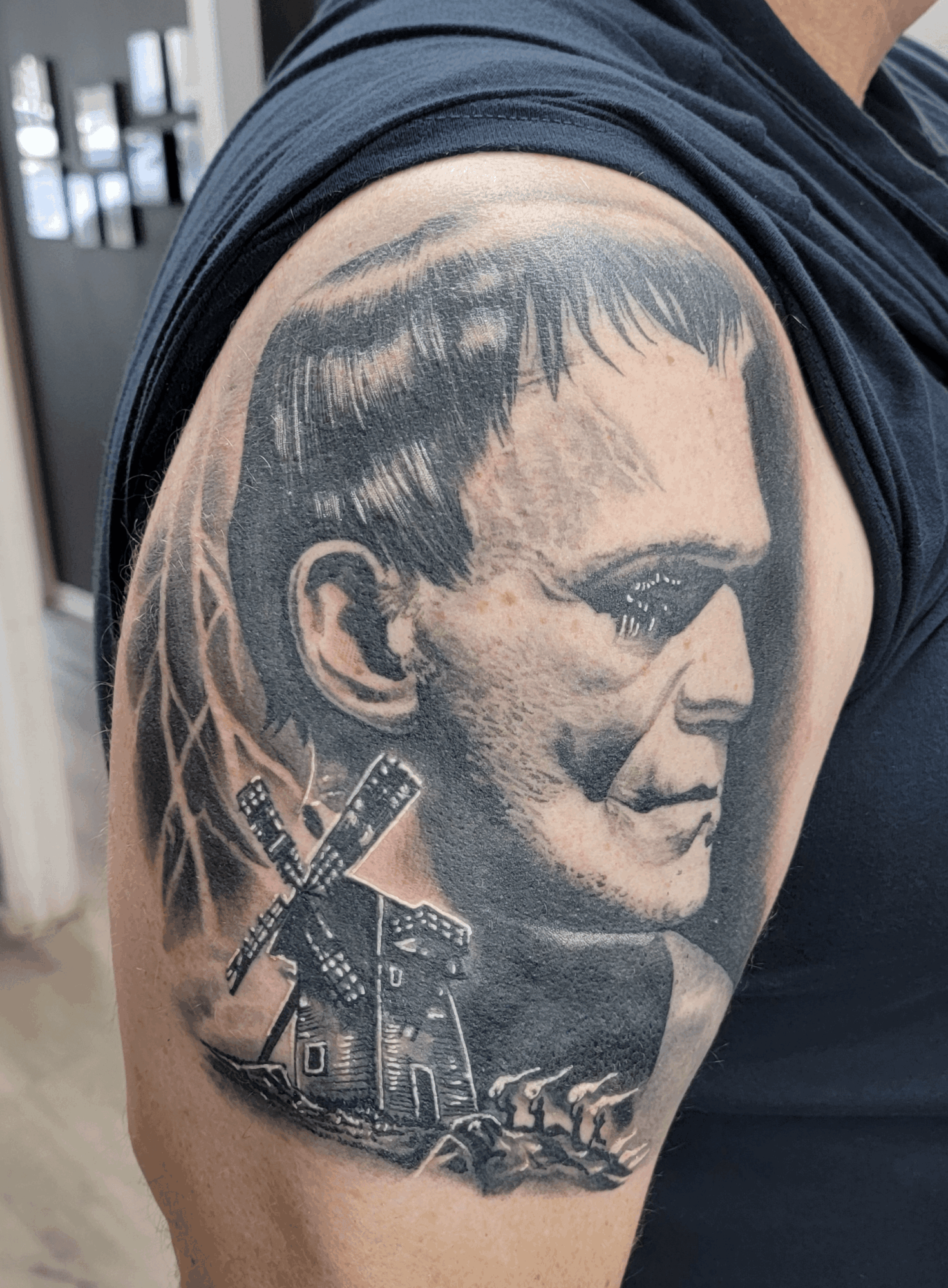 Tattoo of a man's face with windmill and landscape elements on upper arm, detailed shading and realistic style.