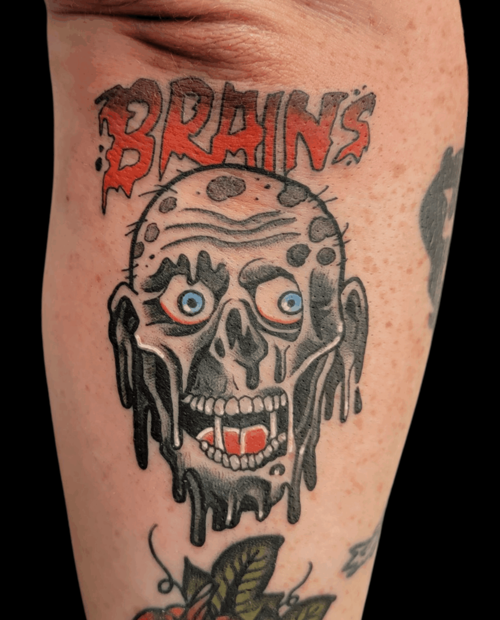 Tattoo of a melting zombie skull with blue eyes and an open mouth, with the word 'BRAIN'S' in red above.