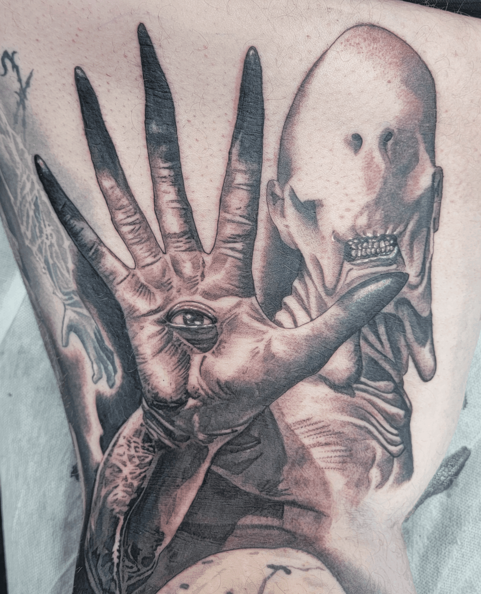 Tattoo of a hand with an eye on the palm and a skull in the background, in black and gray shading.