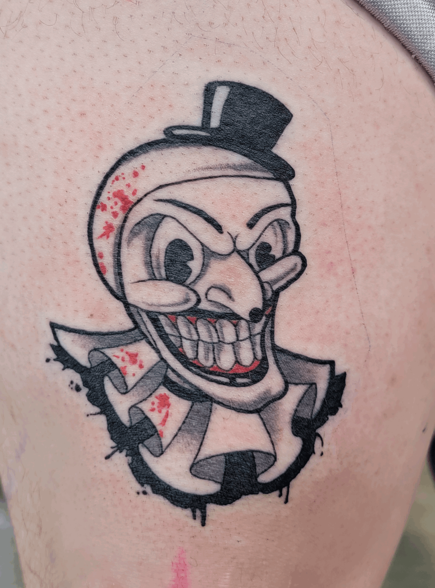Tattoo of a cartoonish skull with a top hat, exaggerated facial features, and a wide grin, on skin with visible pores.