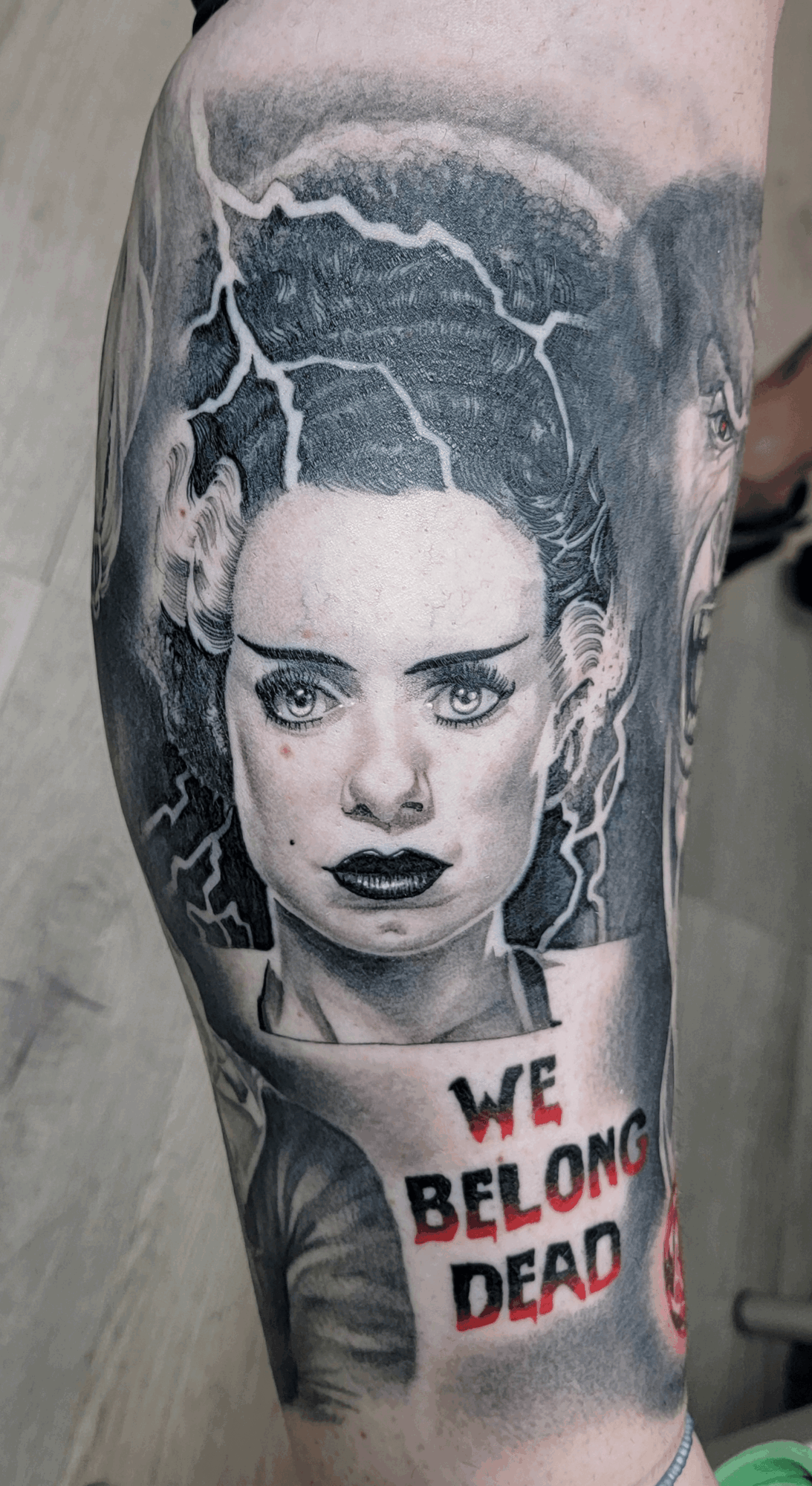 Tattoo of a woman's face with lightning and storm clouds, and text reading 'WE BELONG DEAD' in red and black.