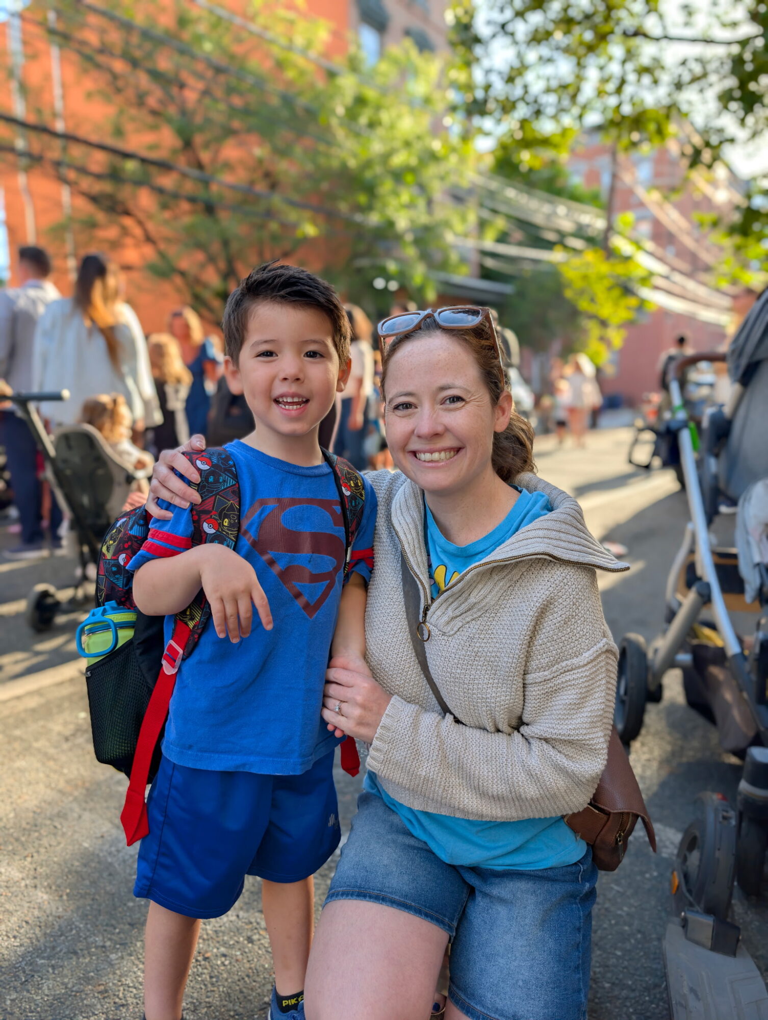 Smiling woman and boy outdoors with trees and people in background, woman wearing sunglasses and hoodie, boy in blue shirt and shorts.