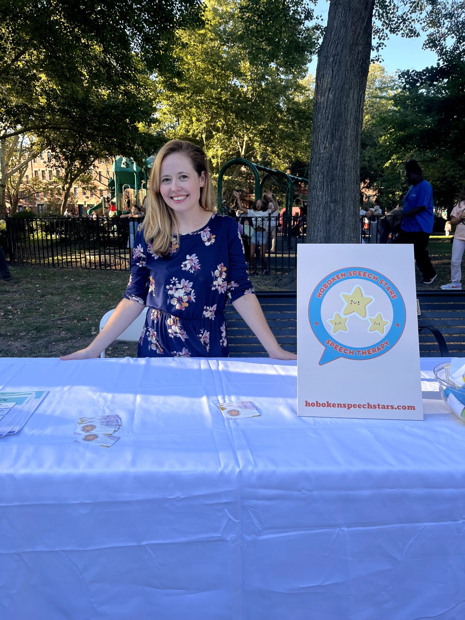 Smiling girl standing behind a table outdoors with trees and people in the background.