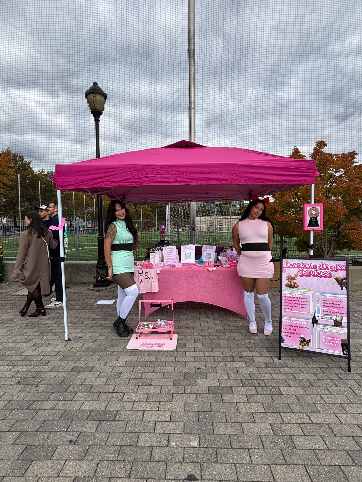 Two women stand at a pink booth with informational posters, under a pink canopy, on a paved outdoor area.