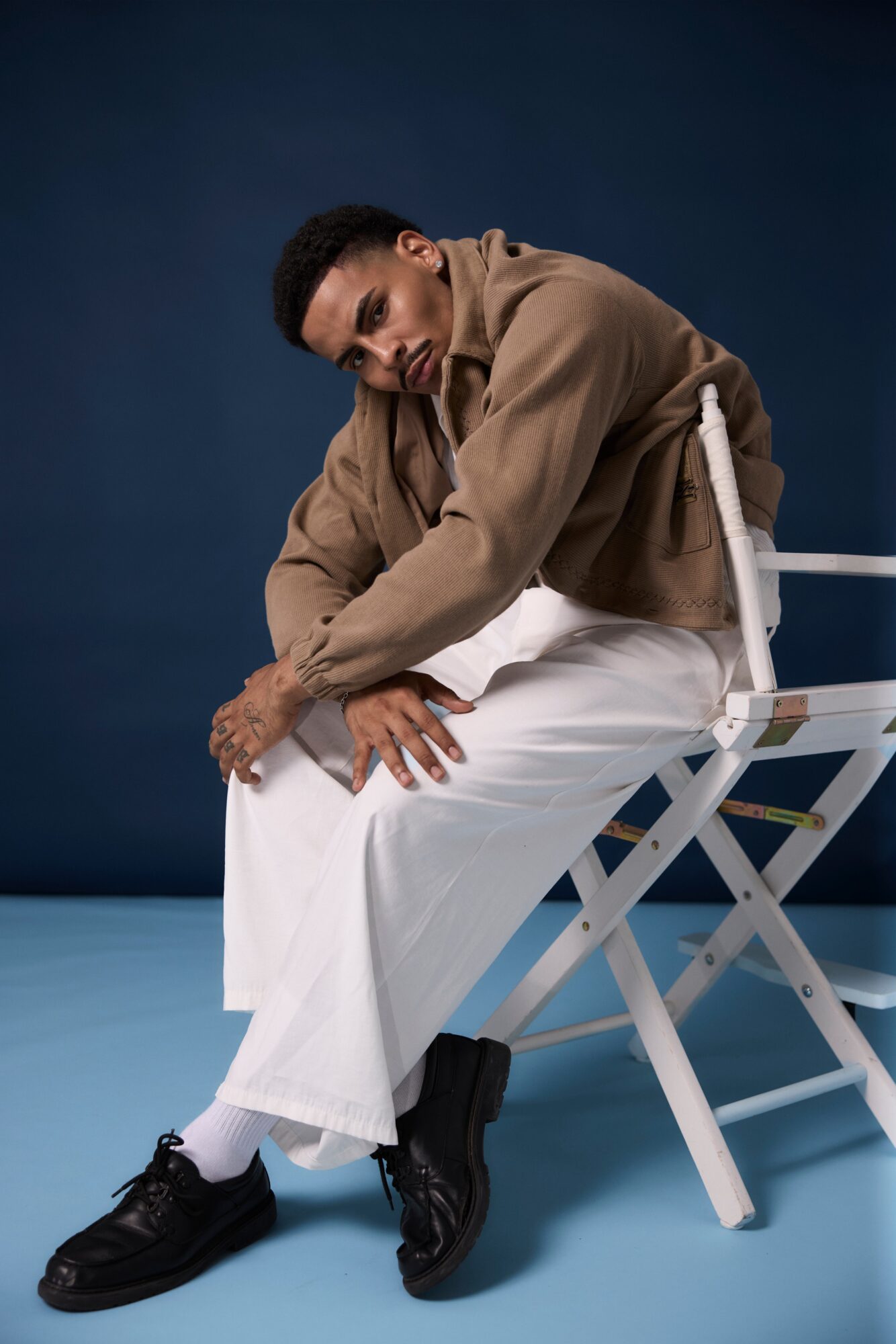 Person sitting on a chair, leaning forward with hands on knees, wearing a brown jacket and white pants, against a dark background.