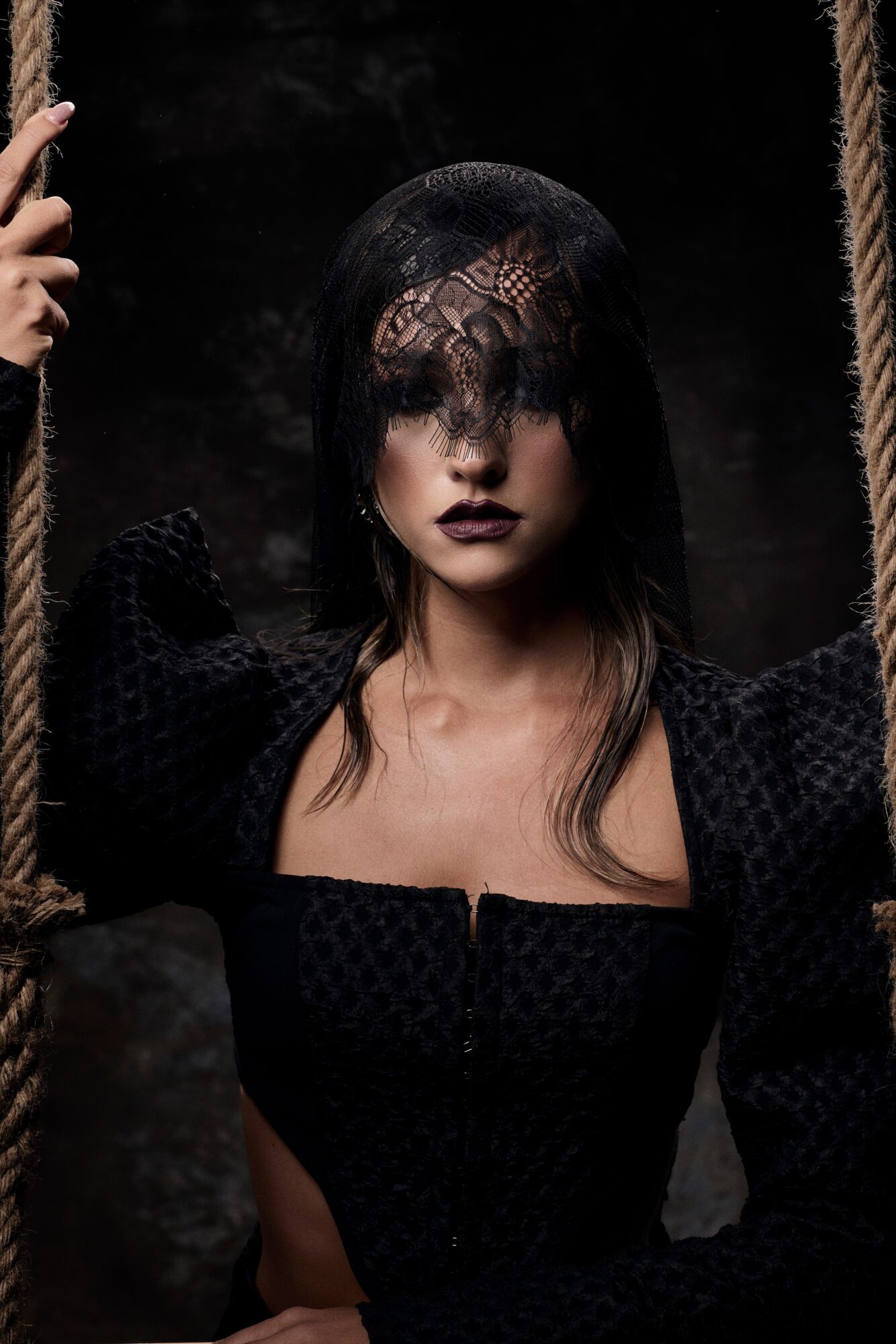 Woman with dark hair and makeup, wearing a black dress, holding a rope, dark background.