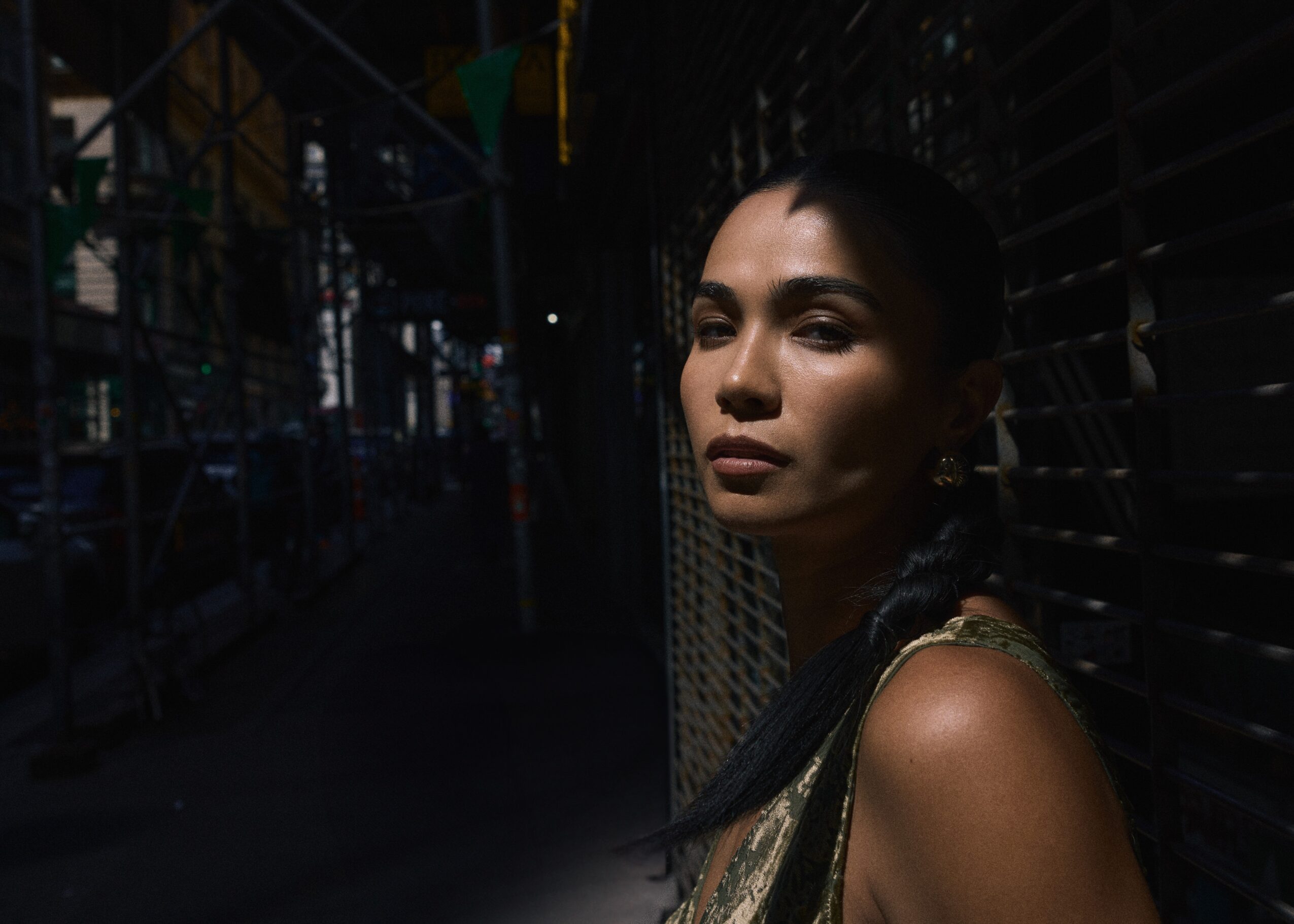 Woman with dark hair and makeup in a dark, industrial setting, looking to the side, illuminated by light on her face.